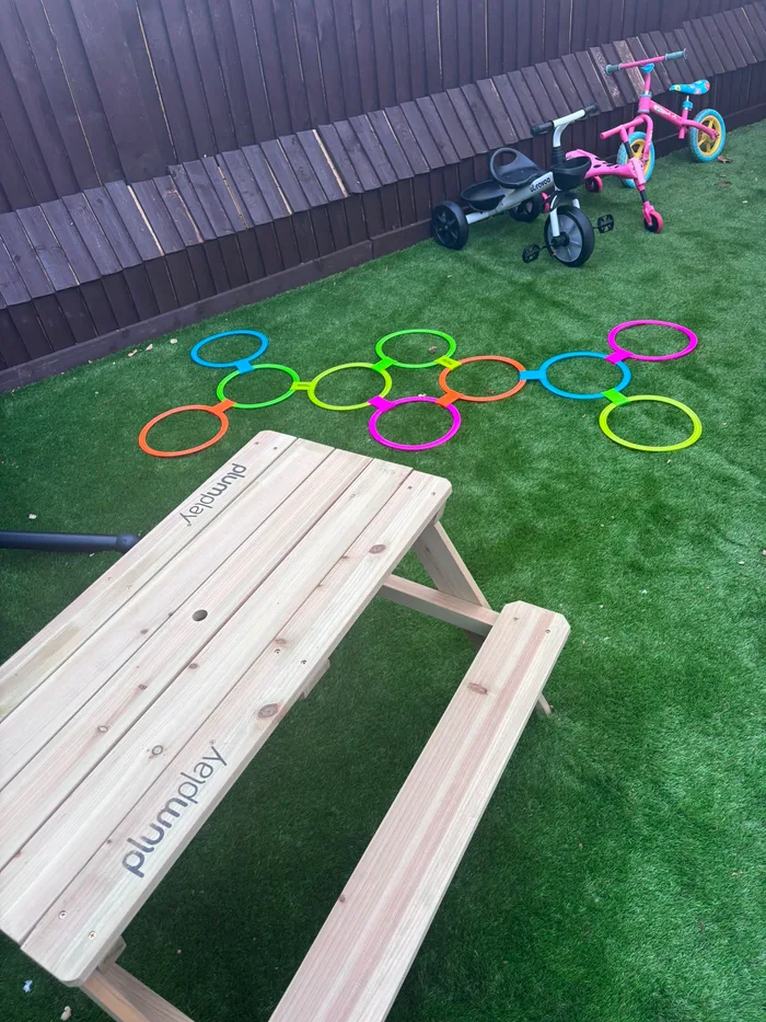 A wooden picnic table on the lawn with a pink trike and hopscotch rings